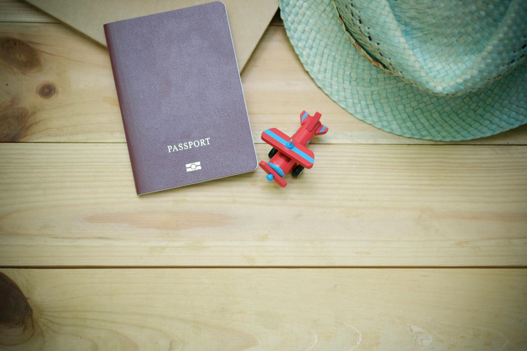 Flat lay of a passport, straw hat, and toy airplane on a wooden surface.